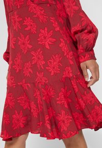 Robe rouge transparente avec des motifs floraux colorés et des manches longues, dotée d'un ourlet évasé à volants et de poignets boutonnés.
