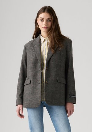 Jeune femme aux cheveux longs portant un blazer gris à carreaux sur une chemise rayée et un jean bleu, debout devant un fond uni.