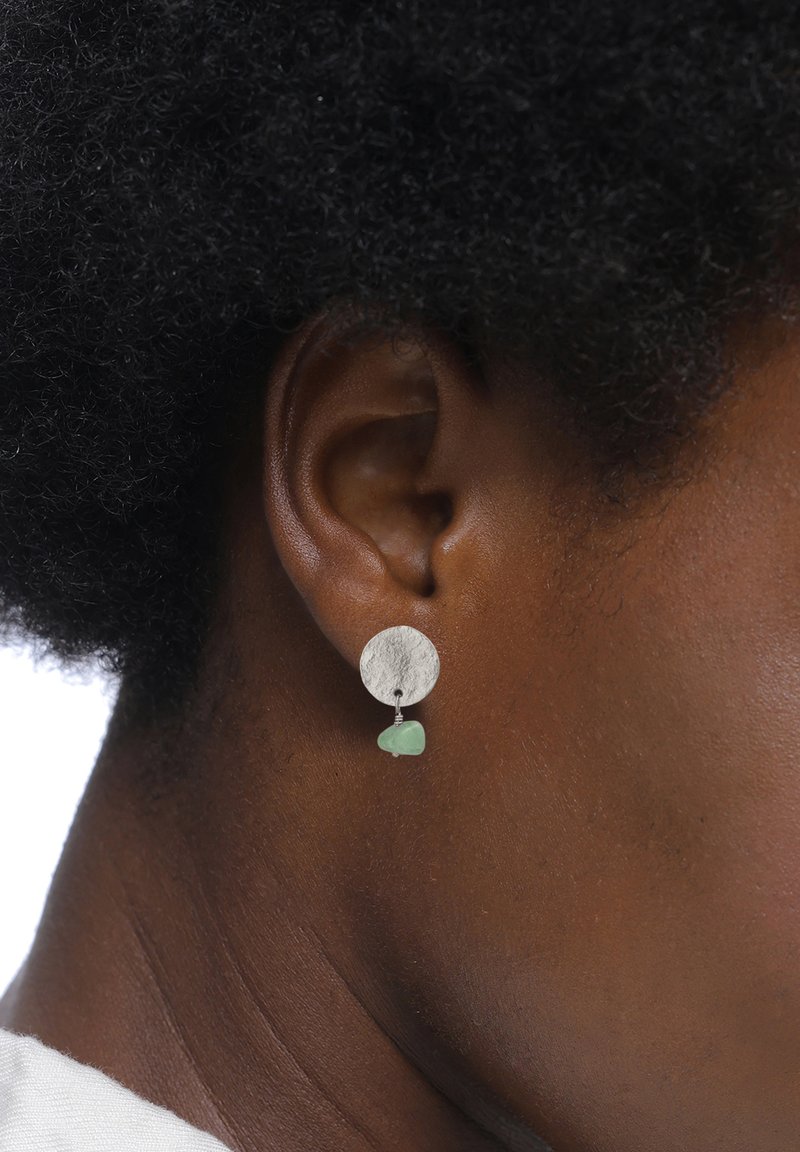Des boucles d'oreilles en argent présentent un disque circulaire texturé avec un accent en verre de mer vert suspendu. Les boucles d'oreilles ont un design simple et contemporain.