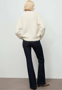 Suéter blanco y esponjoso con mangas largas, combinado con jeans de campana azul oscuro y tacones beige; vista desde la parte de atrás contra un fondo neutro.