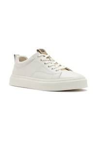 Baskets en cuir blanc avec une semelle en caoutchouc texturé, des lacets plats et un accent beige au niveau du talon arborant un logo. Lignes épurées et design minimal.