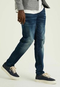 Sötétkék farmernadrág slim fit szabásban, diszkrét kopásokkal, öt zsebes kialakítással és használt hatással. Tengerészkék sportcipővel és szürke dzsekivel párosítva.
