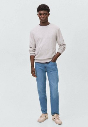 Felpa grigia chiara con polsini a coste, jeans di denim blu e sneakers beige. Il modello sta dritto con una mano in tasca. Design minimalista.