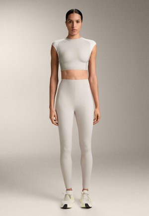 Vrouw staand, naar voren gericht, gekleed in een lichtgrijze cropped sporttop, bijpassende hooggetailleerde legging en witte sneakers met gele accenten.