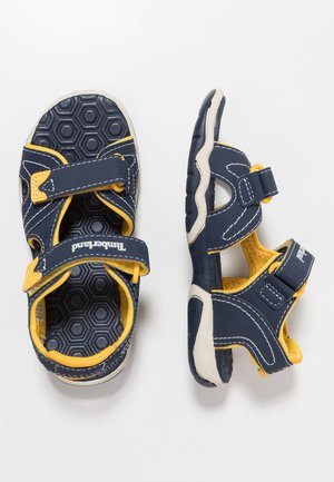 Kinder-Sandalen in Marineblau und Gelb mit sechseckig strukturierten Sohlen und verstellbaren Klettverschlüssen, gezeigt von oben und von der Seite.