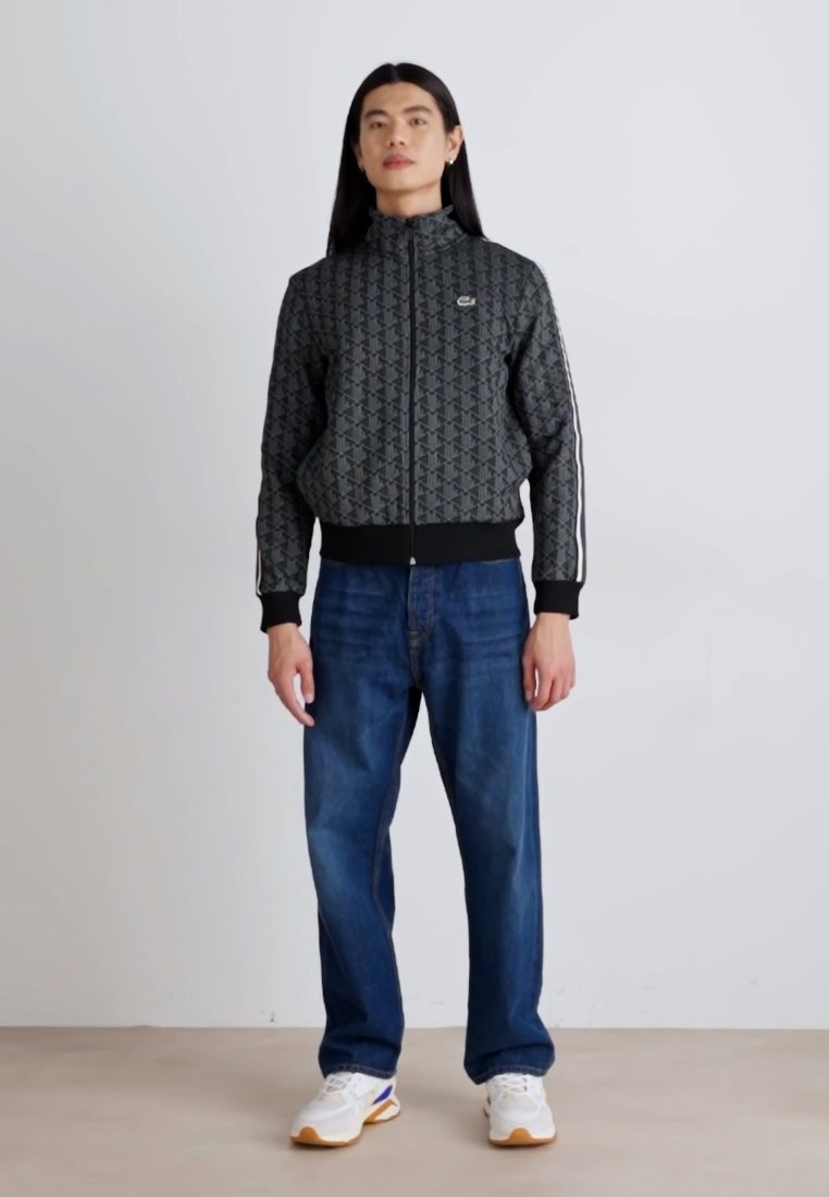 Veste bomber noire à motif avec un col haut, fermeture éclair et bandes latérales, associée à un jean bleu et des baskets blanches.