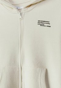 Sweat à capuche crème zippé avec poches avant et petit texte noir sur le côté gauche de la poitrine indiquant « New Futures » ainsi que des phrases motivantes.