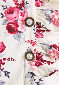 Blouse fleurie présentant des roses roses et rouges sur un fond crème. Fermée solidement avec des boutons vintage en perle et ton or.