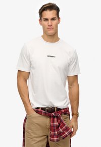 Camiseta blanca de algodón de manga corta, con un logo negro de "SUPERDRY". Pantalones beige con una camisa de franela a cuadros roja y negra atada a la cintura.