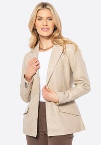 Beige leren blazer met een gestructureerd ontwerp, twee voorkruiden en knoopdetails. Gekleed over een witte top en bruine broeken.