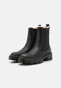 Bottes chelsea en cuir noir avec des panneaux latéraux en élastique, semelles en caoutchouc épaisses, et une doublure douce de couleur claire. Elles sont dotées d'anses à l'arrière.