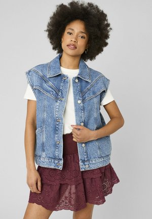 Femme portant une veste en jean bleu clair sans manches sur une chemise blanche et une jupe en dentelle bordeaux superposée, debout devant un fond uni.