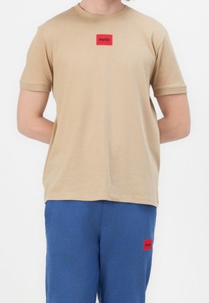 Osoba ubrana w beżowy T-shirt z krótkim rękawem i niebieskie spodnie, oba z małymi czerwonymi prostokątnymi naszywkami marki Hugo.