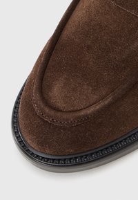 Brown suede slip-on shoe with a rounded toe, stitched accents, and a black rubber sole featuring a textured trim and a smooth finish.