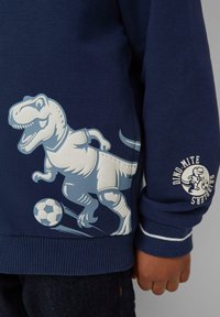 Donkerblauwe sweatshirt met een grote lichtblauwe dinosaurustekening, die voetbal speelt, en de tekst "Dino Mite Dribblers" op de mouw.