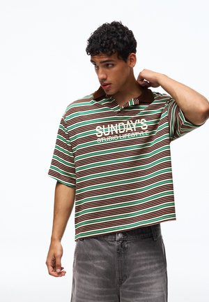 Mladý muž si upravuje golier, má na sebe zelenú, hnedú a bielo prúžkovanú košeľu s nápisom „SUNDAY'S STUDIO LIFESTYLE“ a vyblednuté čierne džínsy.