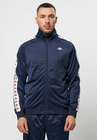 Jachetă de trening bleumarin cu guler înalt, fermoar frontal și dungi laterale albe contrastante, cu un model repetitiv de logo. Textură netedă.