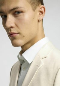 Jasno beżowy dopasowany garnitur o teksturowanym wykończeniu, w połączeniu z elegancką białą koszulą. Posiada spiczasty kołnierz i subtelny detal szwu na ramieniu.