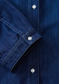 Colletto e maniche di camicia in denim blu, con bottoni bianchi e dettagli di cucitura in evidenza, realizzata in tessuto di cotone texturizzato.