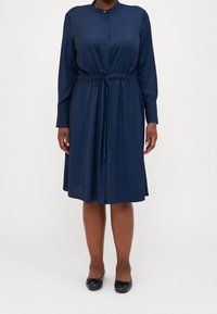 Navyblaues Kleid aus glattem Stoff, mit Kragen, langen Ärmeln, verstellbarem Taillengürtel und knielanger Länge. Schwarze Ballerinas runden den Look ab.