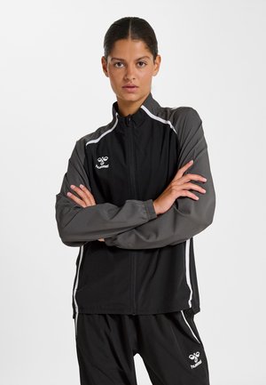 Jonge vrouw met gekruiste armen, gekleed in een zwart-grijs Hummel sportjack en bijpassende broek tegen een witte achtergrond.