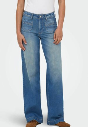 Personne portant un jean bleu à jambes larges avec des poches plaquées à l'avant et des chaussures marron, debout devant un fond clair uni.