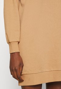 Beige huppari, jossa on ribbed hihansuut ja helma. Pehmeä kangas, rento malli, ja näkyvä käsi, joka lepäilee lähellä alareunaa.
