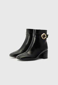 Bottines en cuir noir avec un bout pointu, un talon épais et un accent décoratif de boucle en or sur la sangle. Texture lisse, design minimaliste.