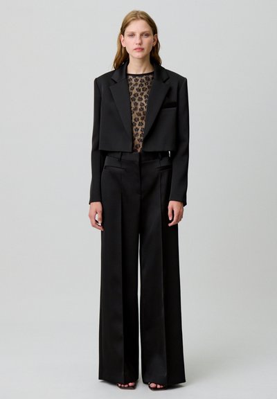 Blazer cropped nero sagomato sopra una maglietta nera trasparente con pattern floreali, abbinato a pantaloni a gamba larga in un tessuto morbido.