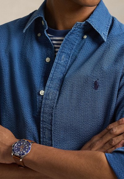 Mees kannab tekstuuriga sinist nööpidega särki sinise ja valge triibulise T-särgi peal, vasakul randmel pruunist nahast rihmaga kella, käed risti.