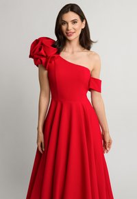 Czerwona wieczorowa suknia z fasonem na jedno ramię, z jedną dużą falbaniastą rękawą, dopasowaną górą i zwiewnym dołem. Gładki materiał o miłej w dotyku fakturze.