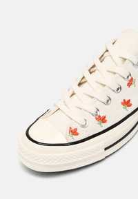 Baskets en toile écrue avec des lacets blancs, une bande noire sur la semelle et une petite broderie de fleur rouge le long du côté près des œillets métalliques.