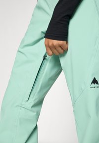 Pantalones impermeables verde menta con un bolsillo lateral con cremallera y detalles de logo. La tela parece suave y ligera, diseñada para uso al aire libre.