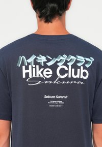 Persona que lleva una camiseta azul marino con el texto "Hike Club Sakura" en inglés y japonés, además de "Sakura Summit" y la dirección del Monte Fuji.