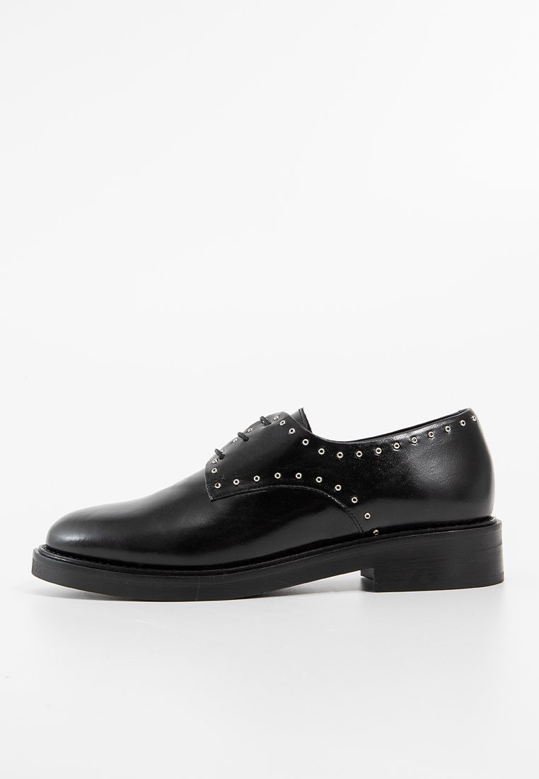 Chaussure derby en cuir noir avec un design épuré, un bout rond et des clous en métal argenté sur les côtés, reposant sur une semelle noire légèrement surélevée.
