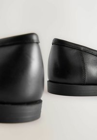 Scarpe in pelle nera con una texture liscia, punta arrotondata, tacco basso e dettagli semplici. La suola presenta un motivo a righe discreto.