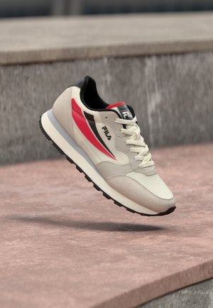 Baskets FILA blanches et beiges avec des accents rouges et noirs, flottant au-dessus d'une surface en béton et d'une marche en extérieur.