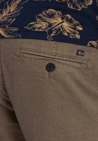 Pantaloni in lino marroni con tasca laterale e bottone nero, abbinati a una maglia blu con motivi floreali dorati. Etichetta del marchio visibile.