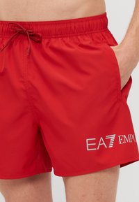 Calções de banho vermelhos feitos de tecido suave. Apresentam uma cintura elástica com cordão e bolsos laterais. Exibem um logótipo prateado na parte frontal.