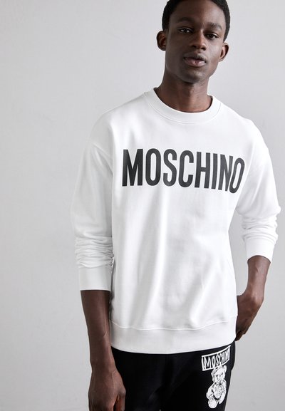 Sudadera blanca con "MOSCHINO" en letras negras en negrita. Con un cuello redondo y puños de canalé. Combinada con pantalones negros que tienen un gráfico de un oso.