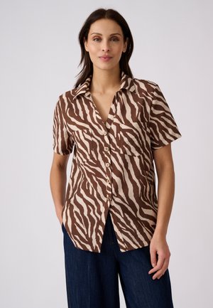 Donna che indossa una camicia marrone e beige a righe zebrate con maniche corte e bottoni e pantaloni blu scuro su sfondo semplice.