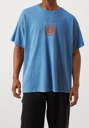 Modré bavlněné tričko s volnějším střihem, kulatým výstřihem a krátkými rukávy. Na přední straně je centrální grafika s nápisem "Escape Reality" a mandalovým motivem.