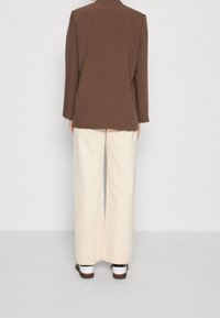 Blazer marrón a medida con hombros estructurados, combinado con pantalones anchos color crema. Ambas prendas presentan texturas suaves y detalles mínimos.