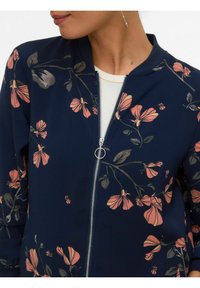 Femme portant une veste zippée bleu marine à imprimé floral rose et un haut blanc, montrant un pendentif de fermeture éclair en forme d'anneau argenté et une boucle d'oreille pendante.