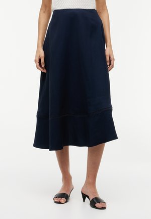 Mujer con una falda midi azul marino con dobladillo acampanado y sandalias negras de tacón abierto, de pie con los brazos relajados a los lados.