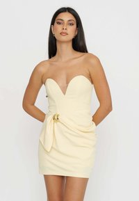 Abito giallo senza spalline con scollatura a V profonda, dettagli arricciati di lato fissati da un accento metallico, texture liscia e silhouette aderente.