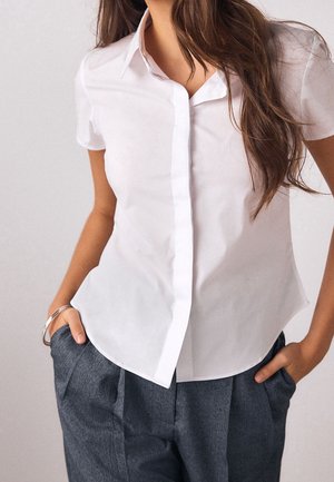 Vrouw draagt een aansluitend wit kort mouw blouse met knopen en grijze geplooide broek met handen in de zakken tegen een effen achtergrond.