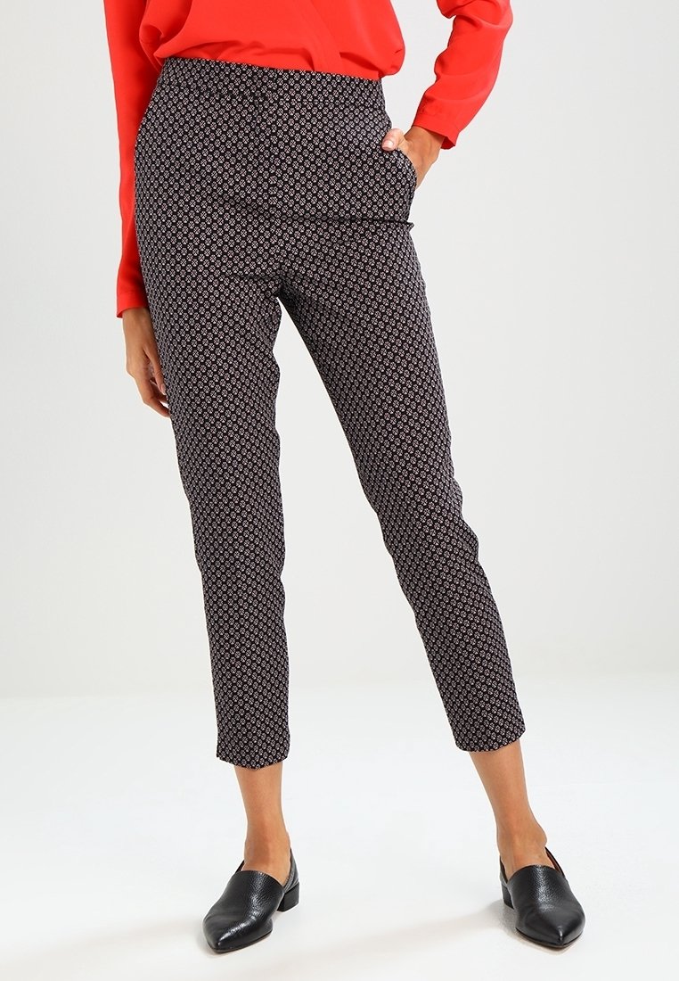 Personne portant un pantalon court noir avec un motif géométrique blanc, des chaussures noires sans lacets et un haut rouge à manches longues avec une main dans la poche.