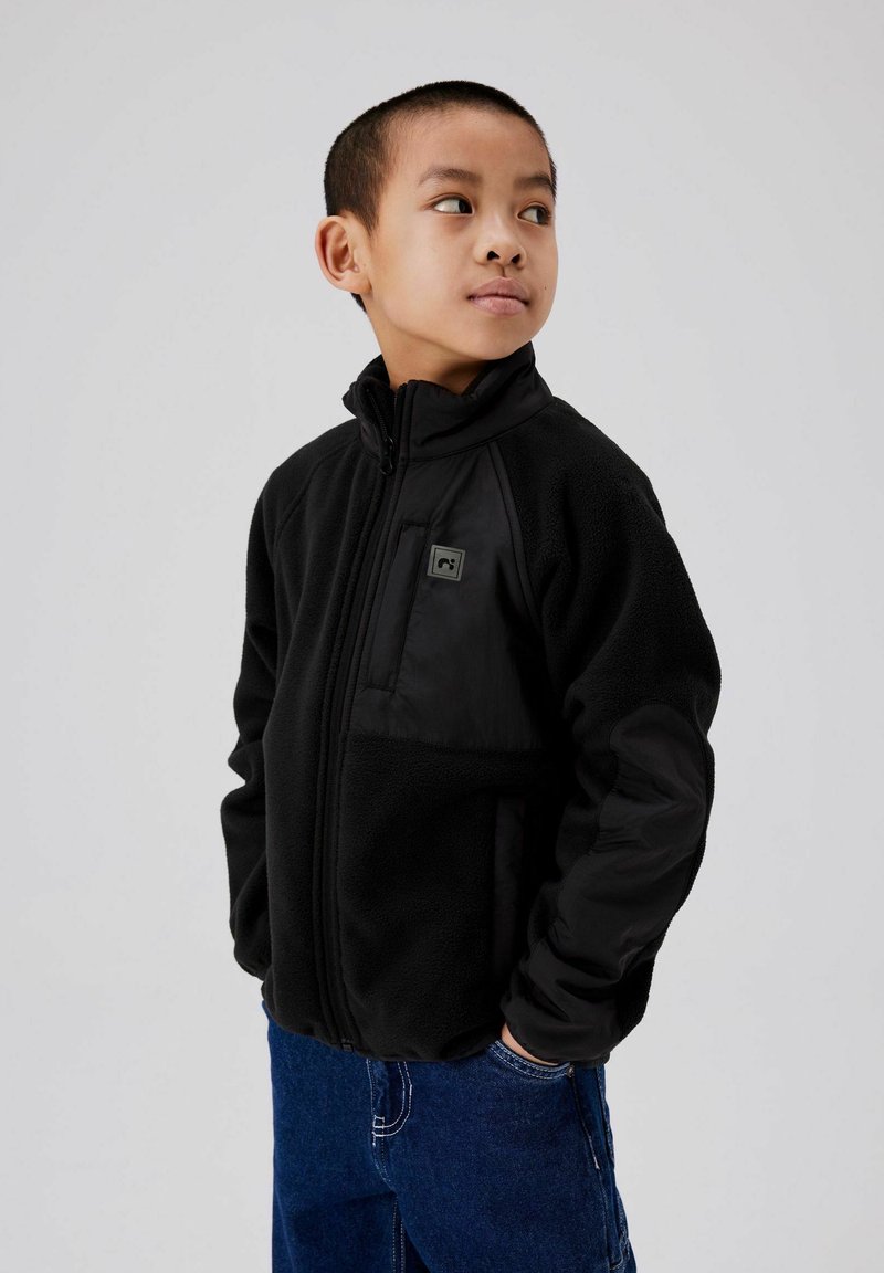 Veste en polaire noire avec un col haut, une fermeture éclair et une poche poitrine avec un logo. Manches lisses avec une texture contrastante.