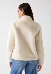 Cremefarbene Kunstfelljacke mit hohem Kragen, lässiger Passform und weicher Textur. Kombiniert mit hellblauen Jeans.